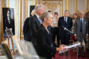 Vorne Premierministerin Elisabeth Borne im Profil am Redepult, im Hintergrund ein Foto von Georges Mandel sowie einige Männer im Anzug