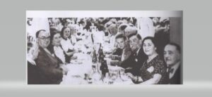 Schwarz-weißes Gruppenfoto, ca 15 Menschen an einem langen Tisch sitzend und in die Kamera schauend