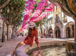 Im Vordergrund ein Brunnen, auf dem eine Frau mit Kleid und Hut sitzt, die nach unten schaut und die Hand ins Wasser hält. Im Hintergrund ein Platz, gesäumt von Bäumen und Häusern, darüber schweben drei Reihen pinker Regenschirme