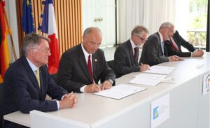 5 Männer in Anzügen sitzen an einem langen Tisch und unterzeichnen Dokumente. im Hintergrund die deutsche, französische und europäische Flagge