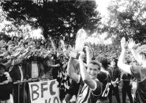schwarz-weißes Foto, Feierlichkeiten nach Gewinn der Meisterschaft, im Vordergrund ein Fußballer, der einen Pokal in die Höhe streckt