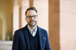 Mann mit dunklen Haaren, Bart und Brille, dunkler Anzug mit dunklem Pullover und hellem Hemd