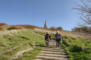 Weg mit Treppen einen Berg hinauf, zwei Menschen gehen die Treppe hinunter, am Gipfel steht eine Kapelle