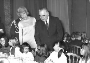 Eine Frau in elegantem Kleid und ein Mann im Anzug, über einen Tisch gebeugt, an dem Kinder sitzen; schwarz-weißes Foto