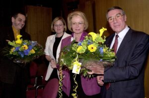Vier schick gekleidete Menschen mit Blumensträußen, in die Kamera guckend