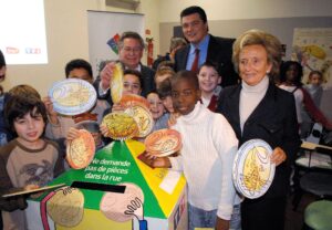 Bernadette Chirac, David Douillet und Dr. Claude Griscelli gemeinsam mit Kindern, die große Euromünzen aus Pappe in einer Spardose werfen