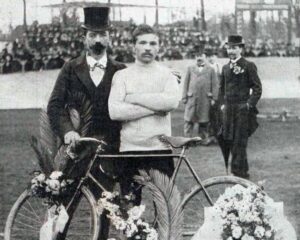 mit Blumen geschmücktes Fahrrad, dahinter zwei Männer, einer mit weißem Oberteil und verschränkten Armen, der andere mit Frack und Zylinder