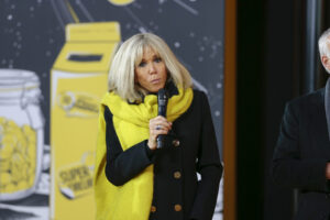 Brigitte Macron mit schwarzem Mantel und gelbem Schal, ein Mikrofon in der Hand