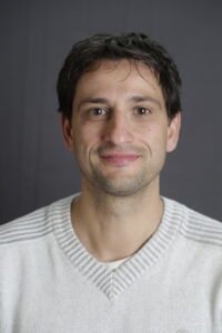 Portrait, Mann mit braunen Haaren und weißem Pullover, leicht lächelnd