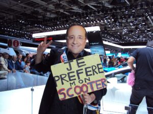 Ein Mensch mit einer Maske mit dem Gesicht von Francois Hollande und einem Schild mit der Aufschrift "Je préfère mon scooter"