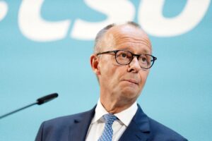 Mann mit wenig Haaren, Brille und verkniffenem Gesichtsausdruck vor einer hellblauen Wand