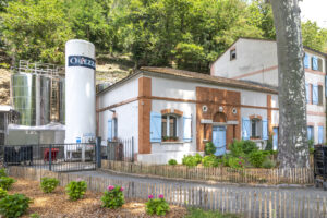 Haus mit heller Fassade und blauen Fensterläden, links davon steht ein großer, runder Wassertank