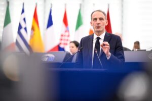 Mann in Anzug und Krawatte an einem Redepult, im Hintergrund verschiedene Nationalflaggen