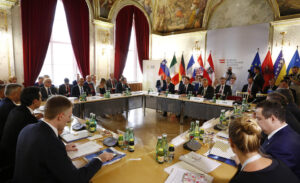 Menschen sitzen an Tischen, die als Quadrat gestellt sind, im Hintergrund Nationalflaggen