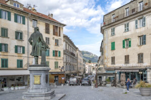 Blick von der Mitte eines Platzes aus, am Ende zwei große Häuser, getrennt durch eine Straße, in der Platzmitte eine Statue
