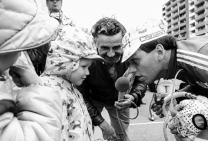 Schwarz-weißes Foto, rechts ein Mann, der sich zu einem Kind (links im Bild) herunterbeugt, im Hintergrund ein Mann, der ein Mikrofon hält