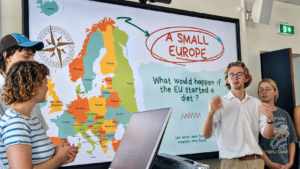 Vier junge Menschen vor einer Leinwand, eine Präsentation mit dem Titel "A Small Europe. What would happen if the EU started a diet" haltend 