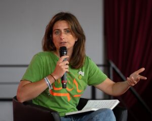 Frau mit grünem Oberteil, sitzend, ein Mikrofon in der Hand