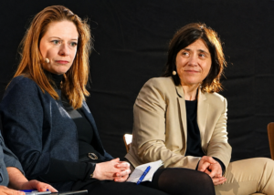 Zwei Frauen sitzend auf einer Bühne, links schulterlange rote Haare, dunkler Blazer, rechts schulterlange braune Haare, heller Blazer