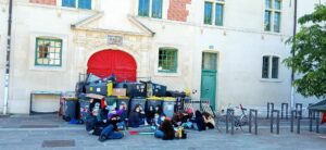 Ein Tor ist mit Mülltonnen blockiert, davor sitzt eine Gruppe Menschen mit Mundnasenschutz, einige schauen in die Kamera