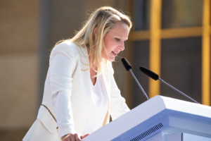 Blonde Frau in weißem Kostüm an einem Redepult, von der Seite abgebildet